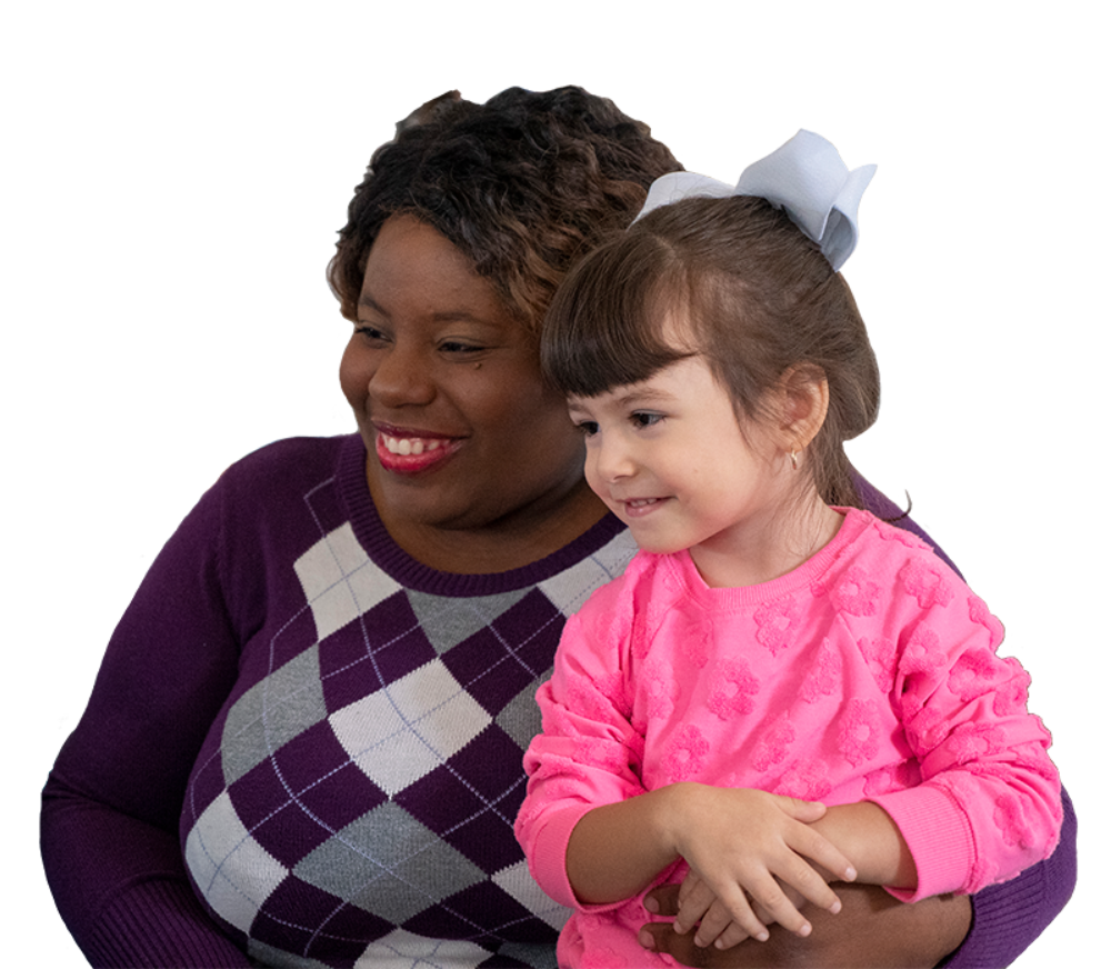 Child care provider in argyle sweater holding a young child in a pink floral sweater with a blue bow.